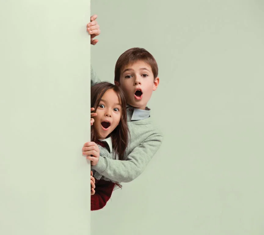Zwei Grundschulkinder schauen neugierig hinter einer Wand hervor